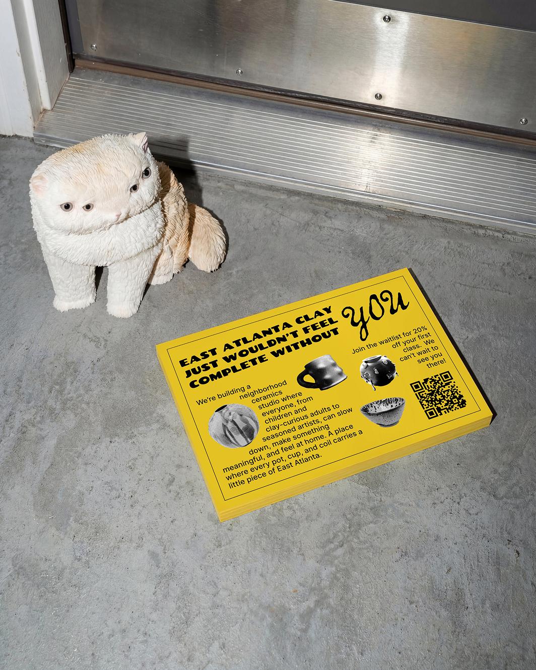 A mockup of a promotional flyer lying in front of a door, with a cat statue that has 4 eyes next to it